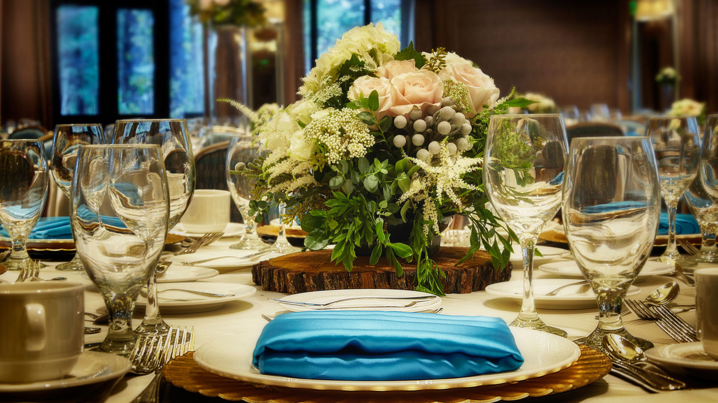 Table set for a reception with a blue silk cloth napkin, glasses, silverware, and a floral centerpiece.