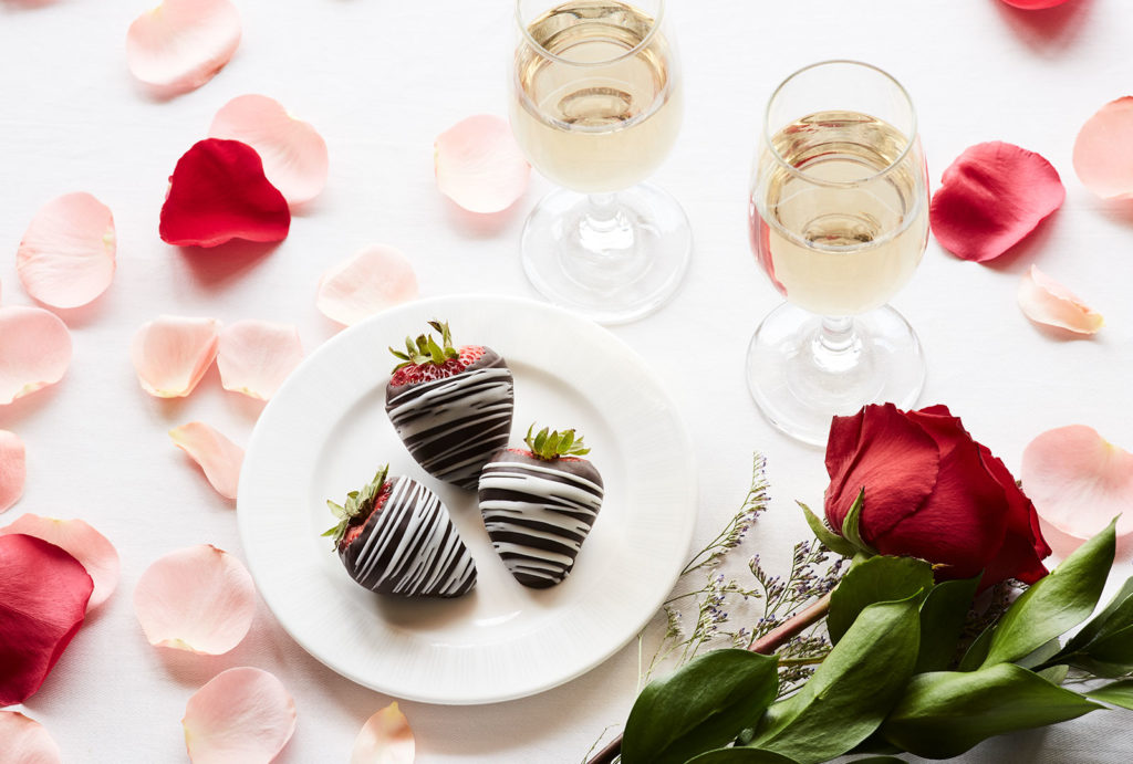 Champagnes and chocolate covered strawberries served with rose petals on a white tablecloth