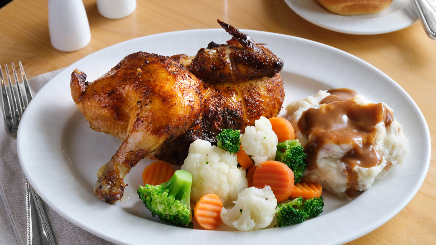 Close up of a rotisserie chicken, mashed potatoes and gravy, and mixed vegetables on a plate.