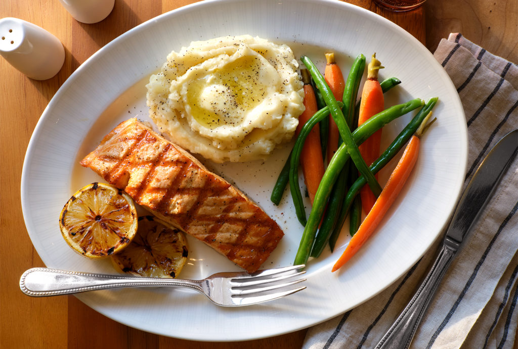 Salmon, mashed potatoes, and beans & carrots on a plate with a fork on a table.