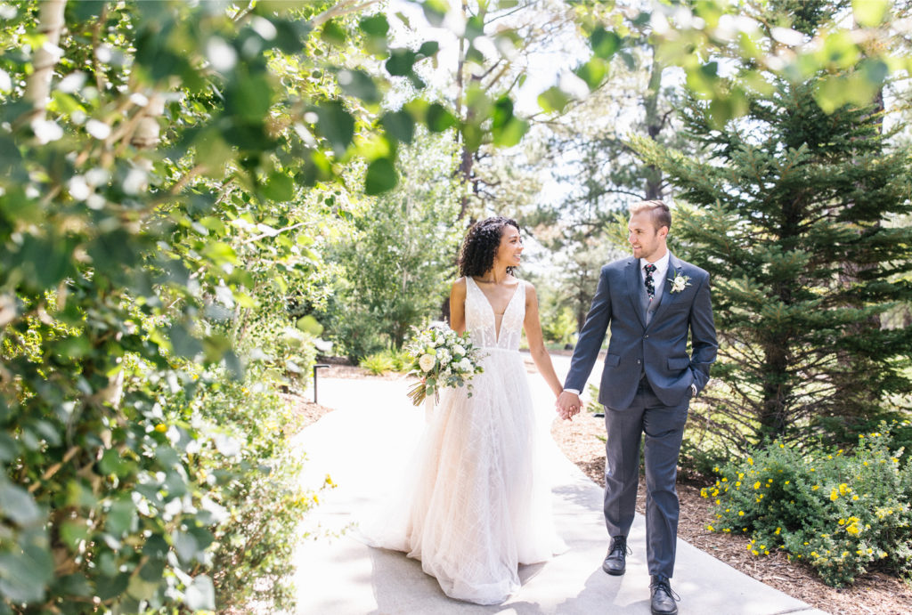 A bride and groom walking together, holding hands on a sidewalk amidst trees.