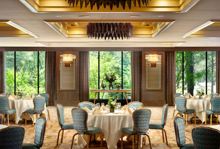 Little America ballroom with tables, chairs, and set tables with three large windows looking into the forrest in the background.